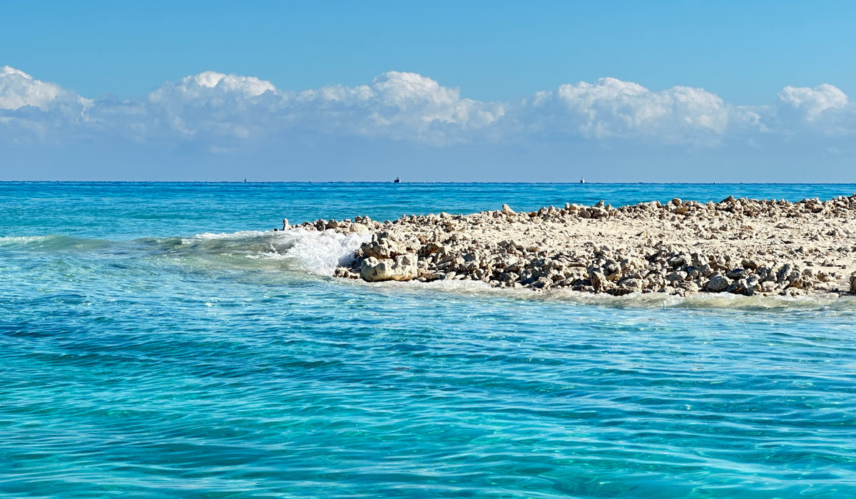 Turquoise Atlantic water breaking over a coral rock spit beneath scattered clouds